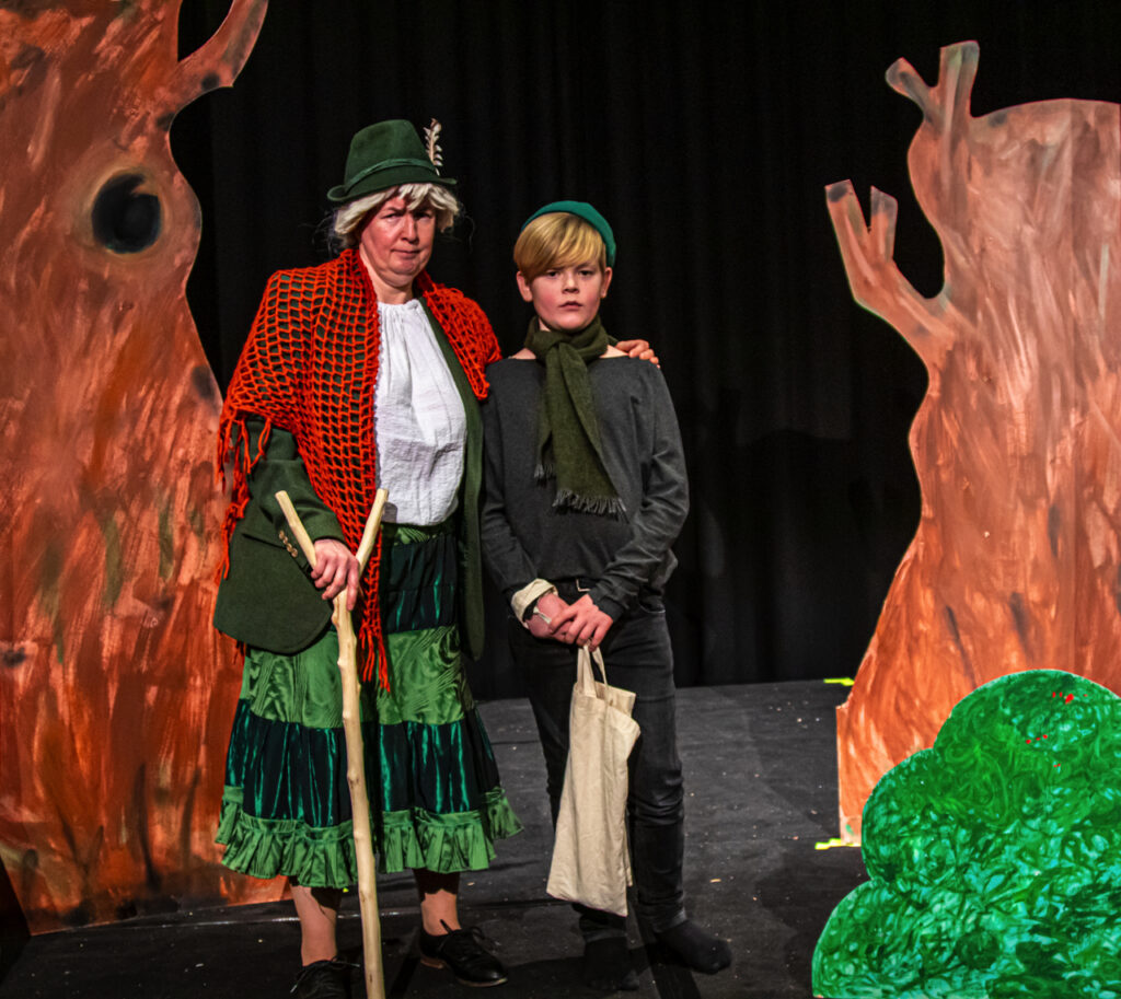 Skuespiller i grønt kostume og dreng i rolle fra Brødrene Løvehjerte, stående på scenen mellem malede træer.
