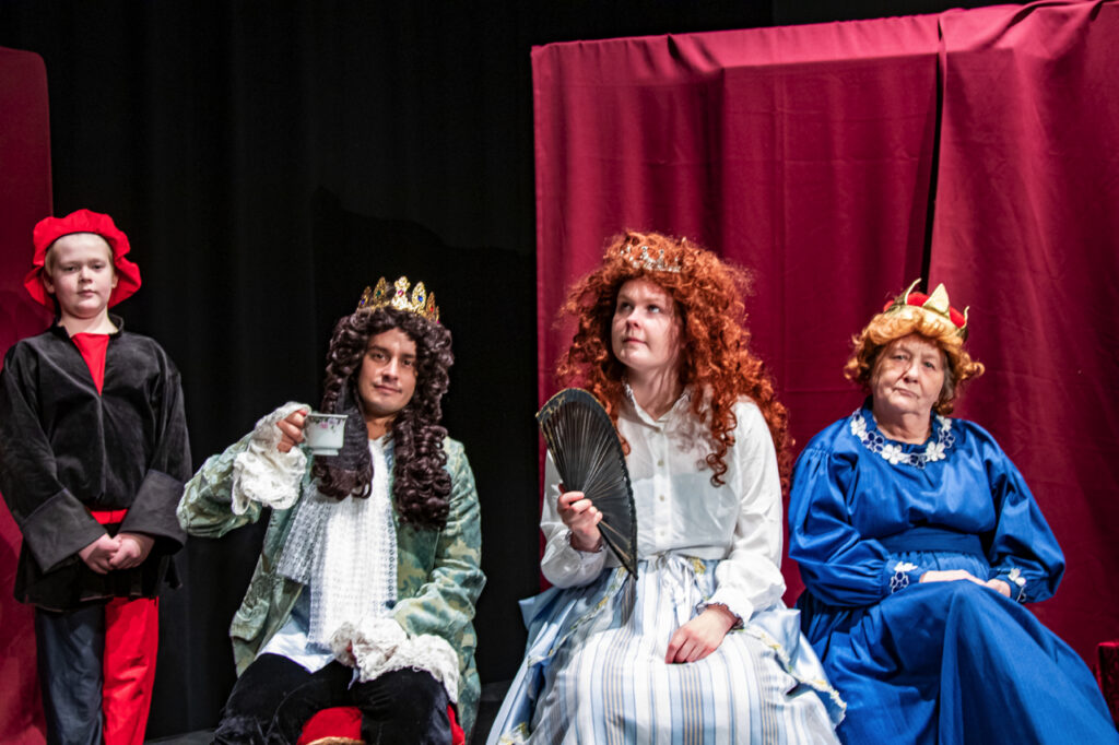 
                        Kongedelige karakterer fra teaterstykket, siddende foran rødt scenetæppe under en forestilling af SkarnTyden