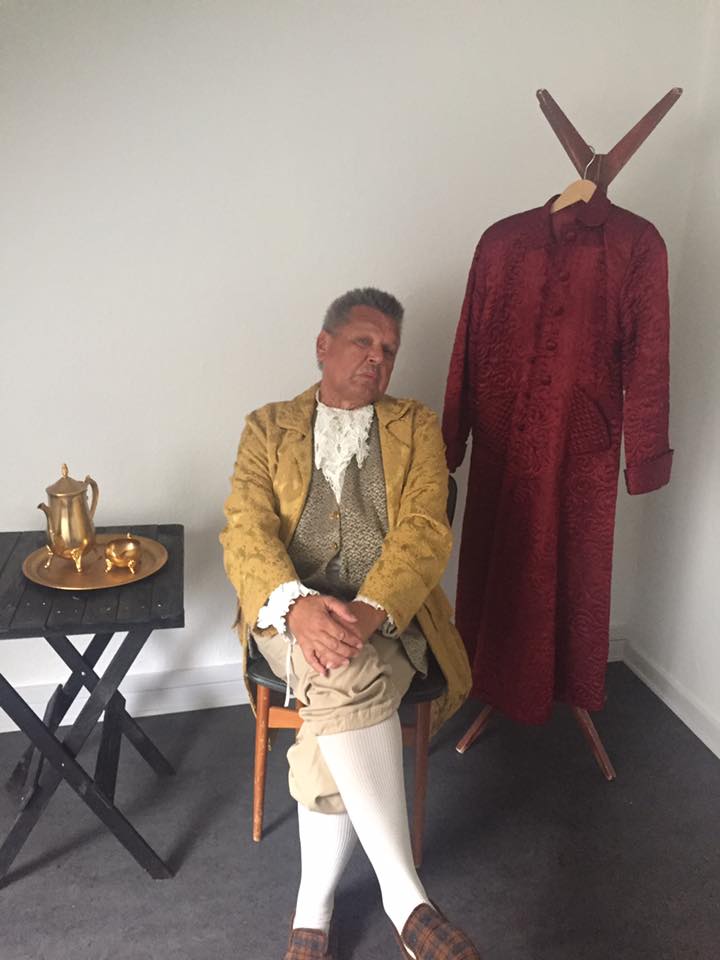 Skuespiller i gult historisk kostume, siddende backstage før forestillingen, ved siden af kostumer og rekvisitter